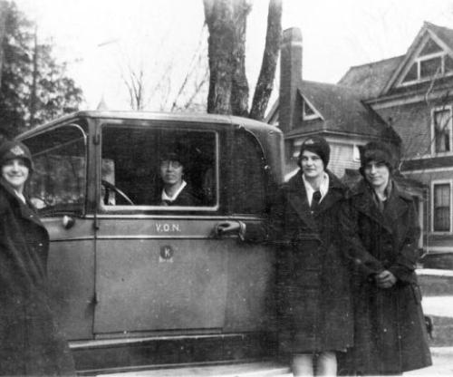 A historic image of three VON nurses in their VON hats and over coats standing infront of car. One nurse in the drivers seat of the car with the window rolled down. circ. 1857
