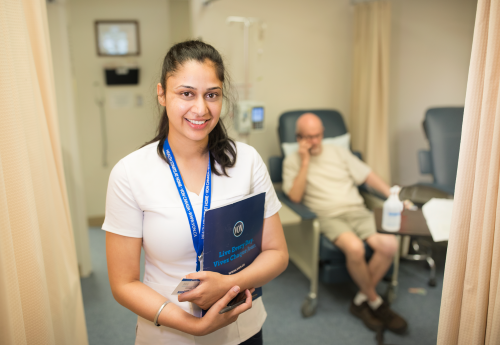 VON nurse looking at the camera smiling with a folder in her arm and standing in the forefront with a client seated in a clinic chair in the background, set in a clinic space. 