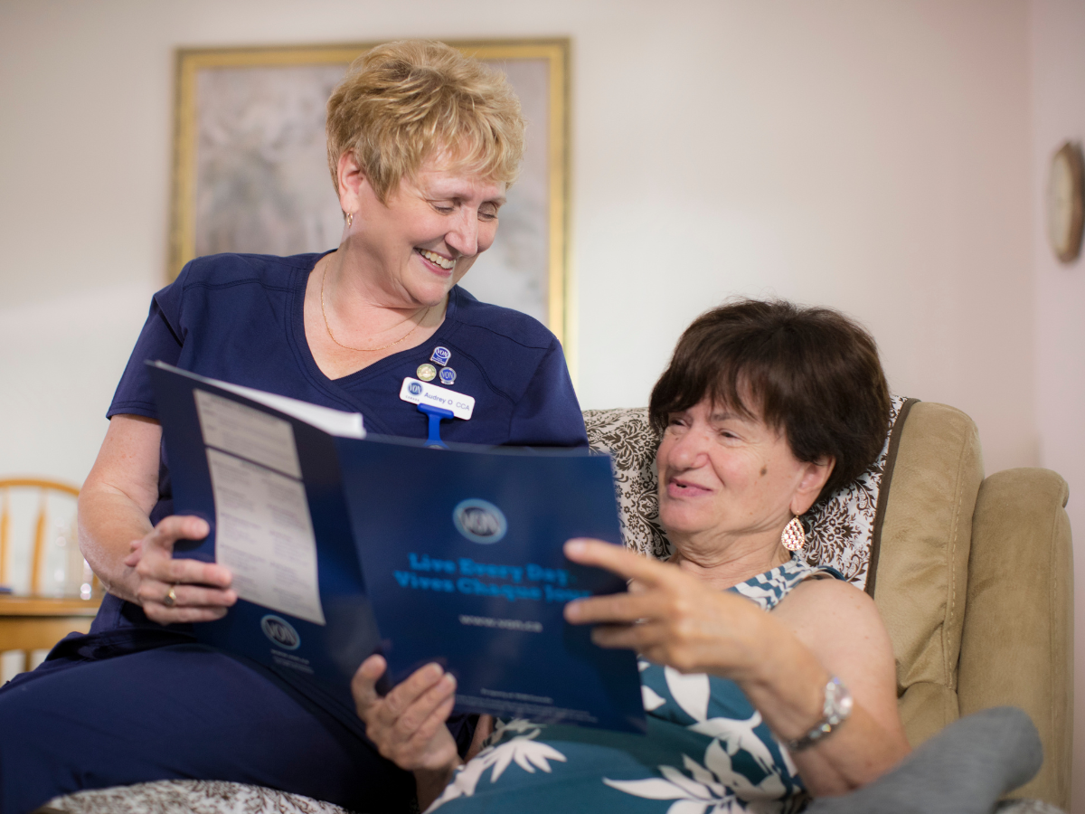 Female staff wearing uniform and sitting on the arm of a chair reviewing paperwork with female client sitting in the armchair.