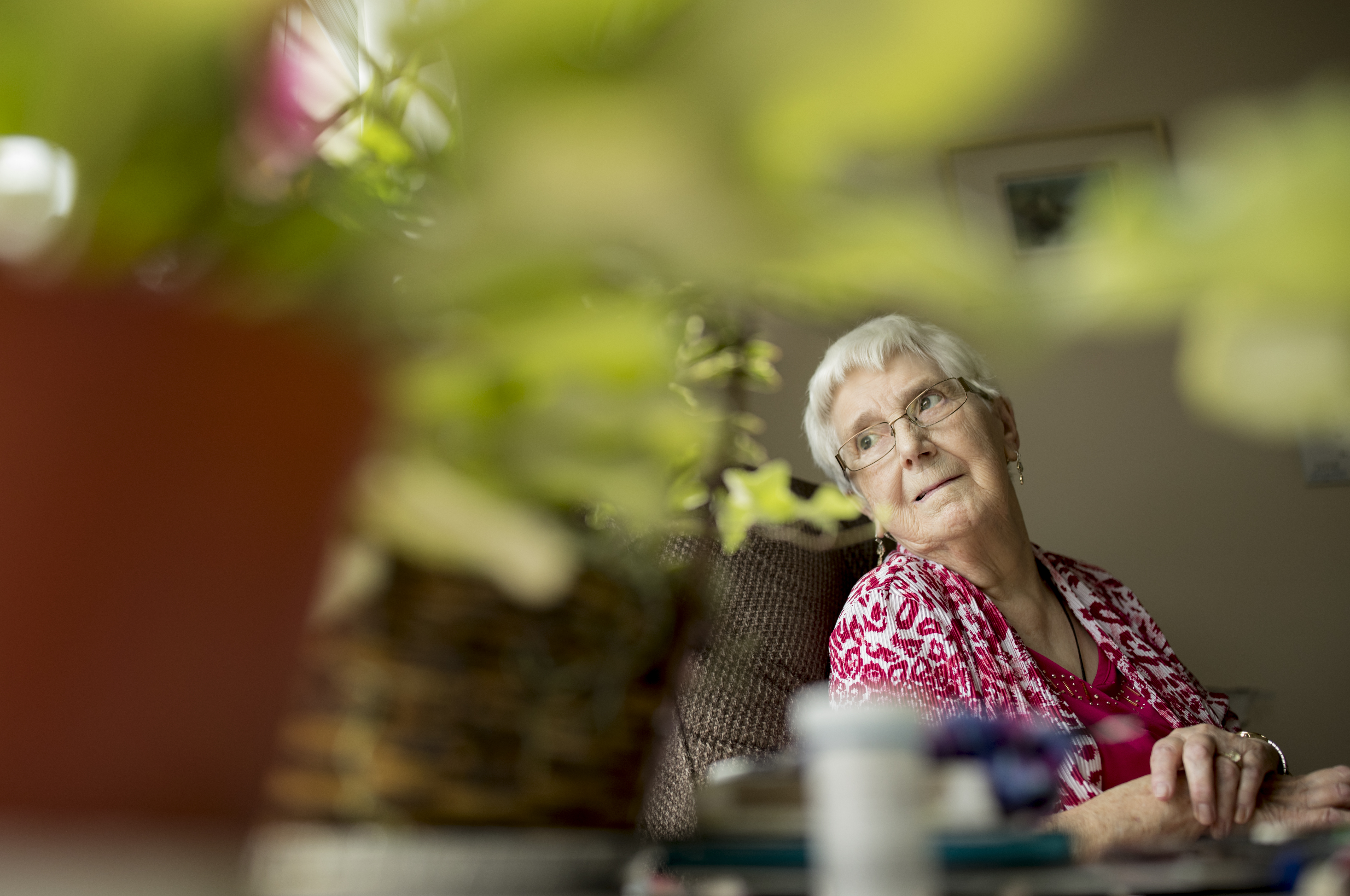 Older female gazing sitting in a chair, gazing over her right shoulder into the distance. A potted plant in the foreground. 