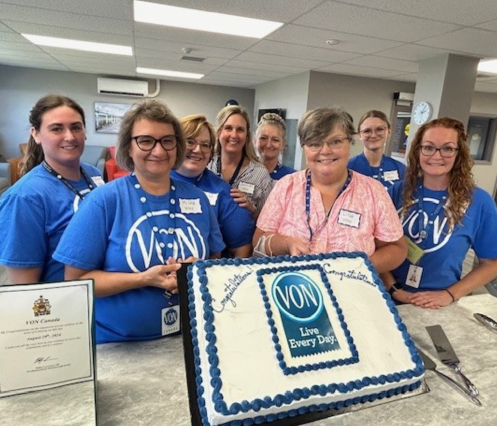 In an office setting, VON staff (women) gather around to pose for a photo with a large cake that has the VON logo with tag that says Live Every Day. on it. 