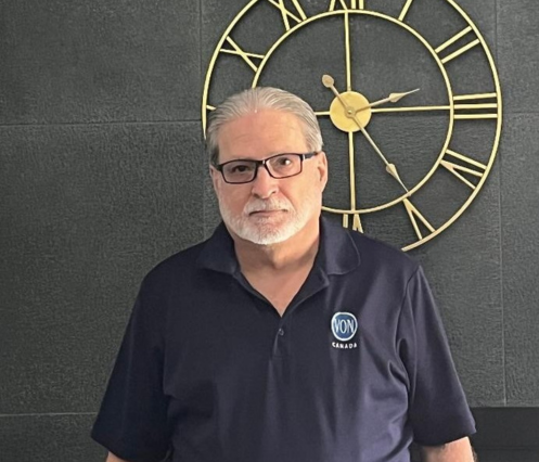 A photo of a man with a white beard and short white beard, wearing black framed glasses and a navy VON polo shirt. Posed in front of a dark wall with a large gold clock.