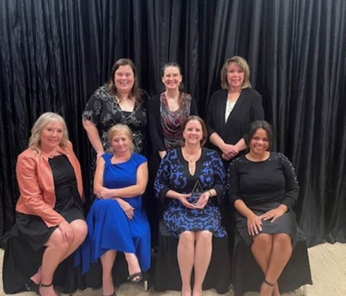 Seven women posed for photo in front of a black curtain. three are standing and four are seated.