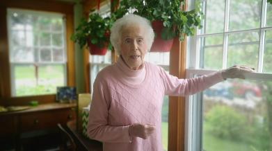 An image of 101 year old Joy Saunders standing in her home by her window, she is wearing a pink sweater. 