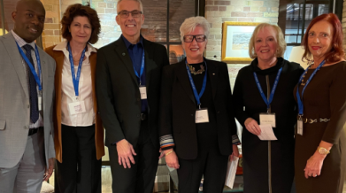 Six individuals posing for a photo, two men and three women, standing indoors in from of a brick wall. 