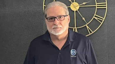 A photo of a man with a white beard and short white beard, wearing black framed glasses and a navy VON polo shirt. Posed in front of a dark wall with a large gold clock.