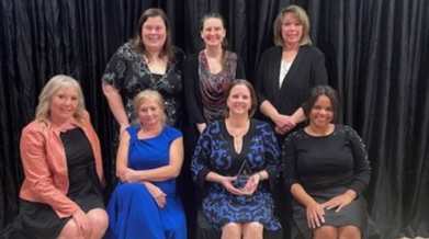 Seven women posed for photo in front of a black curtain. three are standing and four are seated.