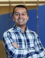 Head shot of Neil Shah, wearing a blue and white plaid collared shirt, arms crossed, looking at the camera. The photo is taken indoors with a blue wall behind him.