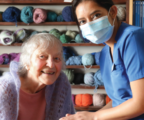 An older female client and a masked VON employee wearing blue scrubs, posing for the photo and holding hands in the clients home. The backdrop is multiple shelves holding skeins of wool.