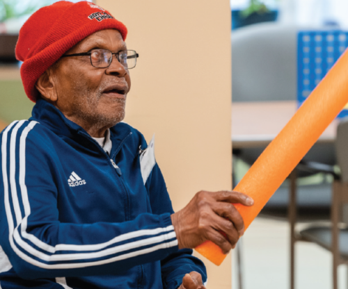 a seated Adult Day Program client wearing a red hat and blue track suite, playing a game with an orange pool noodle inside.