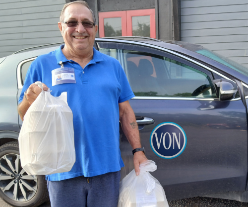 A male volunteer holding a grocery bag of meals, standing in front of VON car